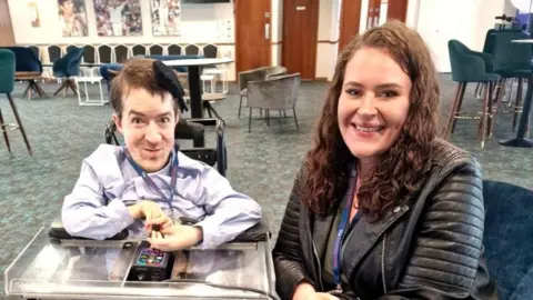 George Baker George Baker and Lauren Toovey smile at the camera. George is on the left in a powered wheelchair, sitting beside Lauren who is wearing a leather jacket in a spacious lounge area with chairs and tables in the background