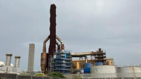 The Guernsey Electricity power plant is in the foreground, with a brown spiral shape and grey cylinders below it. A grey sky is in the background