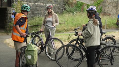 Four people on a cycle path standing holding bicyles while wearing bike helmets. Instructor Andy Smith, wearing a high vis vest, jackets and beige shorts, is standing on the left pointing towards a bridge underpass.
