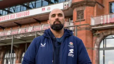 Handout Kabir Ali, wearing a dark blue Lancashire County cricket jacket, poses in front of an empty stand, He has a shaved head and thick neatly trimmed black beard.