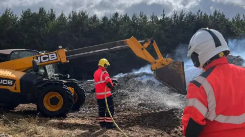 Cambridgeshire Fire and Rescue Service It is daylight and the mound of muck is steaming. There is a yellow JCB digger over the top of it. Two firefighters are looking at the pile of muck facing away from the camera. One is using a hose to spray it with water.