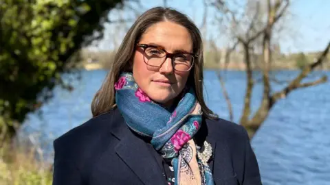 Headshot of Cassie, who is looking at the camera with a serious expression. She has long, straight, dark hair and black-rimmed glasses. She is wearing a navy jacket and colourful scarf, and is pictured outdoors on a sunny day with a river behind her.