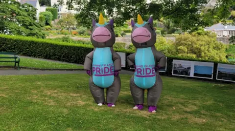 Liberate Two inflatable donkey unicorns, with a blue belly, grey skin, pink nose smiling and a yellow unicorn horn, on the stomach saying 'CHANNEL ISLANDS PRIDE' in a garden under a tree in the shade on a sunny day