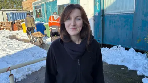 A woman with long dark hair, wearing a black top looks to the camera. Behind her there is snow on the ground, with two people pulling wheelbarrows.