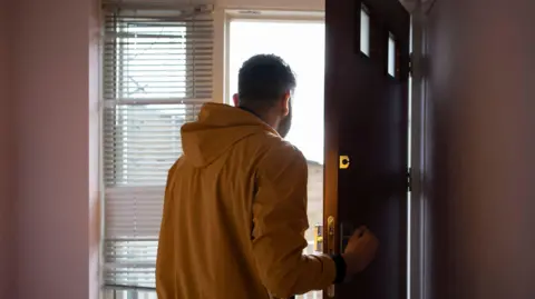 A man in a yellow jacket stood inside opening his front door. The door is brown and there are shutter blinds hanging from the window to the left. The walls are painted a light pink. 