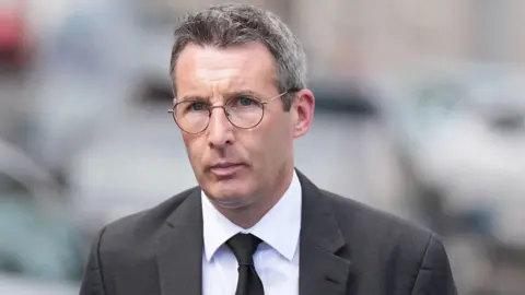 A man with glasses and short dark, greying hair stares into the camera. He is wearing a grey suit, white shirt and dark tie.