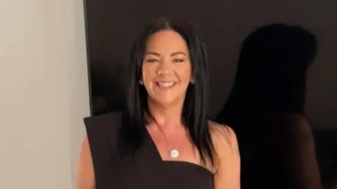 Karen Cummings, who has long dark hair parted in the middle, smiles while wearing a black dress and silver necklace and standing in front of a large flat screen TV