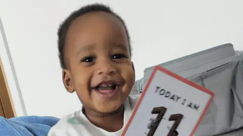 Family handout A smiling baby with short black afro hair is holding a car that reads 'Today I am 11 months old'