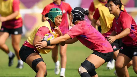 Action Images/Reuters Women playing rugby, dressed in pink tops and black shorts. The player holding the ball is wearing a yellow bib and green helmet. 