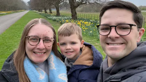 PA Media Jennifer and Matthew, stand with their son William between them, posing for a selfie. All three wear dark coats; Jennifer is wearing a baby blue scarf, has her brown hair down and is wearing glasses. Matthew is also wearing glasses and has short dark hair. William has fair hair. They are smiling at the camera.
