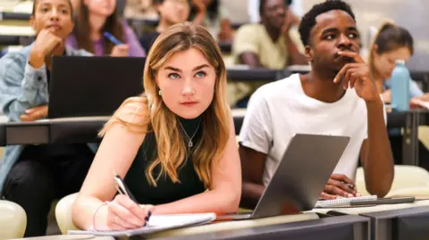 A stock photo of students in a classroom, with one woman jotting in her notepad 