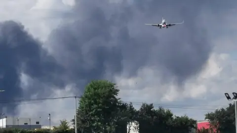 Andy Zetroc A plane flies through billowing black smoke, above a row of trees