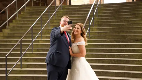 A man in a blue suit with red tie takes a selfie with his wife in her white wedding dress