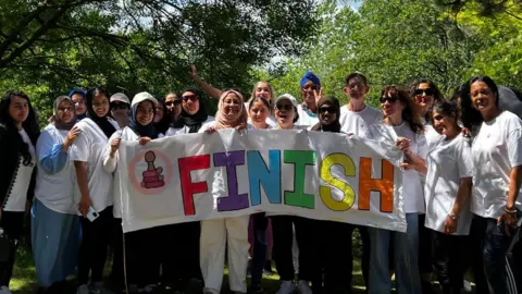 Shantona Women's and Family Services A group of 20 women lined up across the photo. They are holding a sign that reads "finish", and appear to have completed a fundraising event. They are all wearing white T-shirts. They are stood with lots of trees in the background behind them.