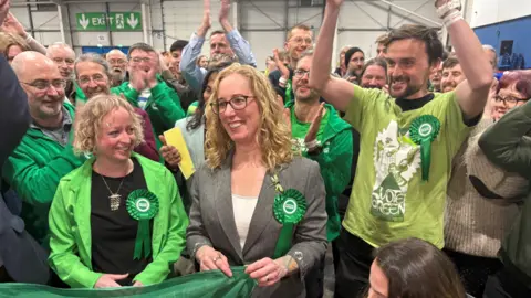 PA Media Lorna Slater, in grey suit and white shirt, is surrounded by Green Party supporters, largely in green. They look very happy. They are in a modern industrial-looking hall.