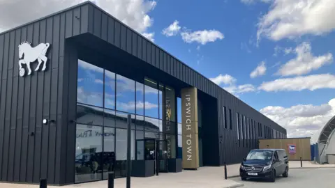 Graeme McLoughlin/BBC A large black building with glass windows and doors. On the right of the building is a black Mercedes vehicle. The words Ipswich Town are on their side running down the side of the entrance, and the club's horse and football logo is on another side of the building.
