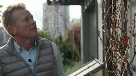 Ed Howden in a shirt and gilet looking at a vine on the outside of his home.