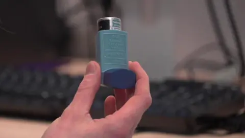 A doctor holds up a blue asthma inhaler. A computer keyboard is in the background.
