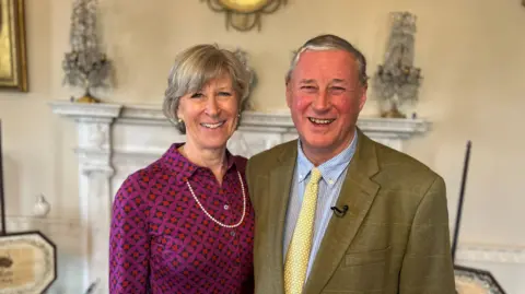 Beth Parsons/BBC News A woman with short hair wearing a purple and red blouse with a single string of pearls arm around a man wearing a green and brown jacket with a blue shirt and pale yellow tie