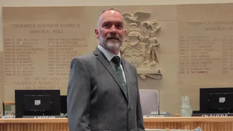 Man wearing a grey suit and tie stands in a council chamber. Behind him are desks and on the wall is inscribed names of previous Thurrock mayors.