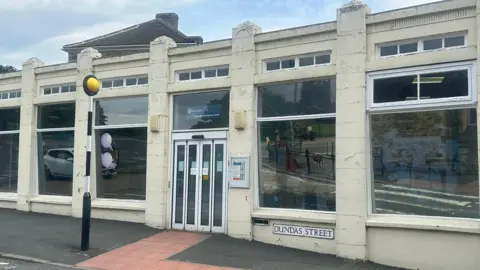 The current library, a white, flat-roofed building with white automatic doors, with a sign saying 'Dundas Street' next to a zebra crossing light.