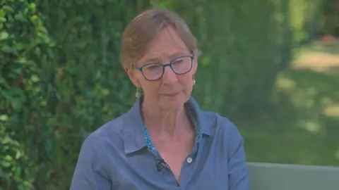 a woman with short fair hair, wearing a blue shirt and glasses is sitting in a gard