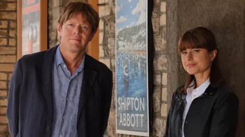 A promotional photo of Beyond Paradise featuring actors Kris Marshall and Zahra Ahmadi. They are standing outside an old building with a sign saying "Shipton Abbot", the setting of the show. Kris Marshall has light brown hair and is wearing a dark navy blazer over a light blue button-up shirt and light beige trousers. He has hands behind his back. Zahra Ahmadi is standing in a doorway, leaning lightly against the wall. She has long dark hair and is wearing a dark, zip-up jacket over a white collared shirt. Her hands are in front of her.