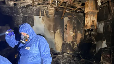 University Hospital Southampton A man in blue overalls and wearing a protective mask is shining a torch in a room that has been completely blackened and destroyed by fire. 