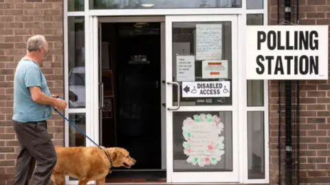 A man in a T'shirt and brown trousers walks with his dog to the entrance of a polling station. The entrance has a polling station sign and other stickers in its glass windows.
