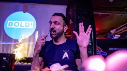 Sid Akbar/Bradford Arts Centre A man puts a fork of food to his mouth with one hand and with the other he holds his fingers in a Star Trek symbol