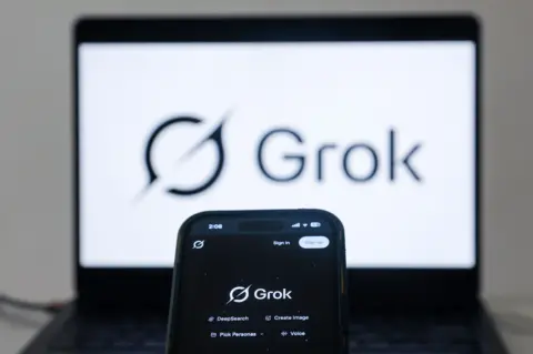 EPA A phone is in the foreground showing a black screen with the word Grok on it. Behind it is a laptop with a white screen showing the same name and a circle logo with a line going through it. 