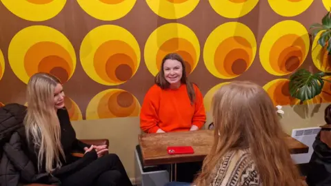 Supplied Three women sitting at a coffee table with snazzy wallpaper in the background. Thale, with her back to the wall, is wearing an orange jumper. She is smiling.