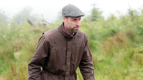 The Prince of Wales walks from grassland while wearing a brown coat and a grey flat cap. He has a trimmed beard.
