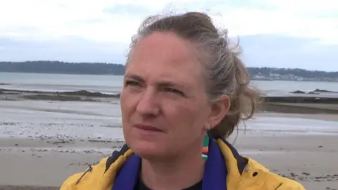 Hilary Jeune is a woman with brown and grey hair staring past the camera. She is wearing a yellow anorak with a purple scarf and a brightly coloured earring in her right ear. She is in front of a beach with sea and a horizon in the background.