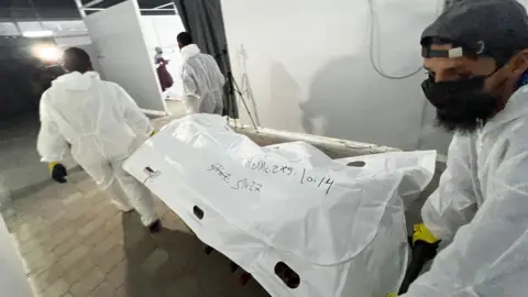 BBC Palestinian officials move a body bag containing the remains of an unidentified Palestinian, which were handed over by Israel as part of the Gaza ceasefire deal, at Nasser hospital, Khan Younis, southern Gaza (14 October 2025)
