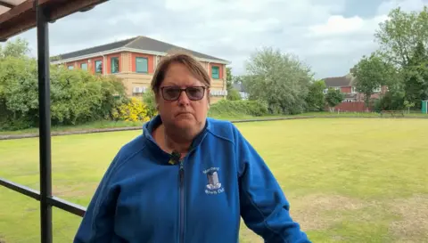 A woman with short brown hair and glasses in a royal blue fleece jacket stands in front of a bowling green.