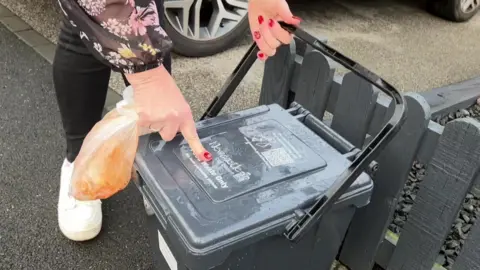 A Newcastle City Council kerbside recycling caddy. A female resident is pulling down the handle to open the caddy and add a bag full of kitchen waste.