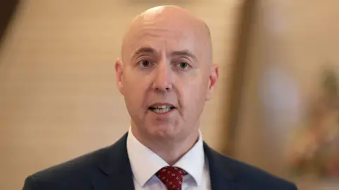 PA Jon Burrows is standing in an empty room. He is wearing a navy blazer, white shirt and red tie 