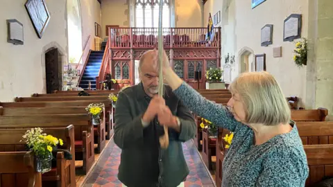 George Carden/BBC Guy Pratt with his teacher in the aisle of the church pulling on a rope connected to a bell in the steeple