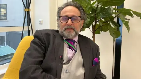 A bearded man with curly mid-length grey hair and wide glasses with black rims looks into the camera. He is sat in a yellow chair, wearing a dark grey suit jacket, silver waistcoat, and a pink and purple pocket square and tie over a pink shirt