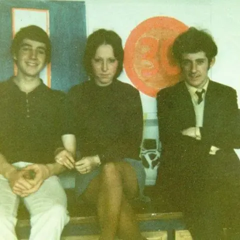 Forever Churchfields Students pictured in the 1960 - a young woman with a short skirt and short brown hair is flanked by two smartly dressed young men, all smiling at the camera 