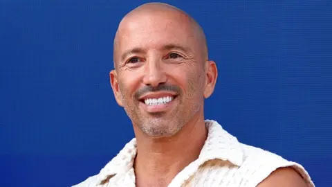 Jason Oppenheim smiling and standing in front of a blue background