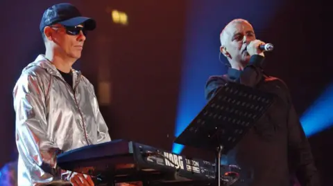 PA Media The Pet Shop Boys on stage in 2022, Chris Lowe, on the left is wearing a silver jacket, cap and glasses as he stands next to a keyboard. Neil Tennant stands close to him holding a microphone to his mouth.
