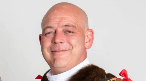 Wolverhampton City Council Greg Brackenridge's head and shouders are pictured. He is looking at the camera in front of an off white background. He is wearing brown fur, a mayoral chain and red robes.