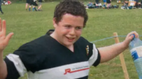 Carl Powles Jak as a teenager with curly brown hair wearing a black and white striped rugby shirt and celebrating with his arms in the air after a win, while standing on a grass pitch.