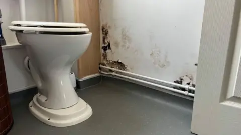 Andrew Turner/BBC Toilet in foreground with evidence of damp damage to plaster on wall and unpainted box work around the sewer stack that leaked