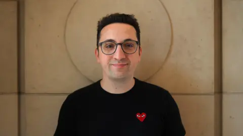 Adam Riches A man with short black hair, glasses, and a black shirt stands in front of a sand-colored wall. His head is perfectly framed by a circular recess carved into the large stone block.