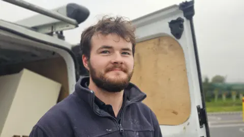 Paul Moseley/BBC Hayden Newport standing wearing a thick, blue works cardigan. He's standing in front of the back of his white trades-van.