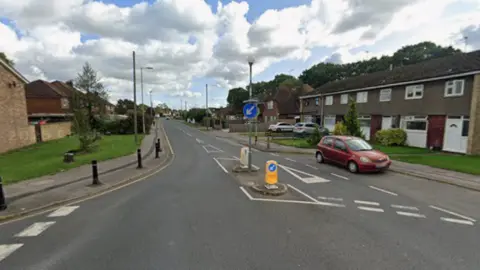 A Google Maps picture of Woodlands Avenue, a long road with houses on either side. A red car is parked to the right of the picture in front of a house and close to a crossing.