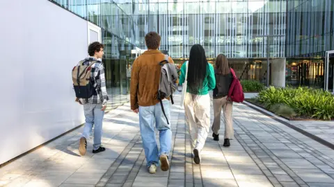 Getty Images Four people with backpacks walk along a paved path towards a modern glass building. The group of students is seen from behind, wearing casual clothes, and the setting includes greenery and reflective windows.